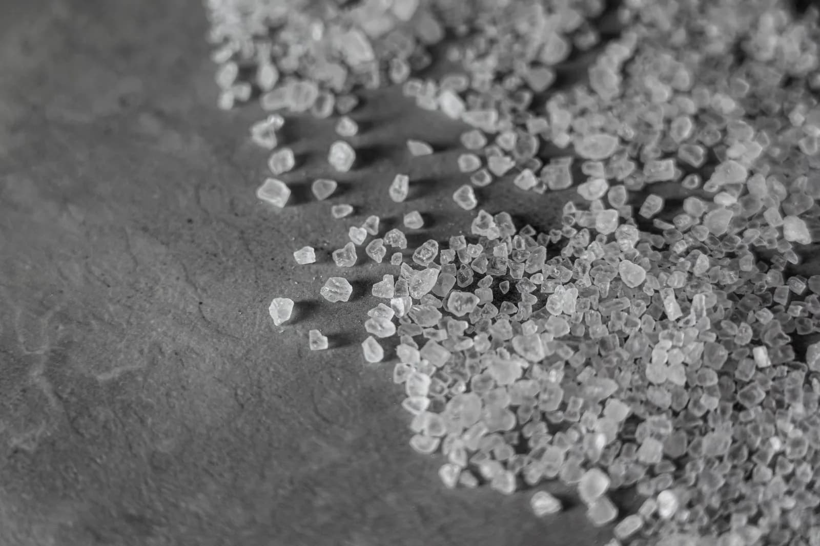 Coarse salt granules scattered across a dark surface, seen close up in black and white