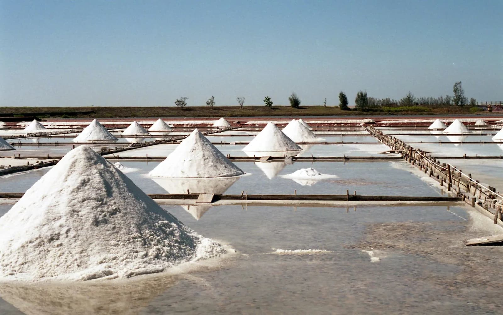 Conical mounds of white salt standing in shallow evaporation ponds divided by wooden walkways under a clear sky
