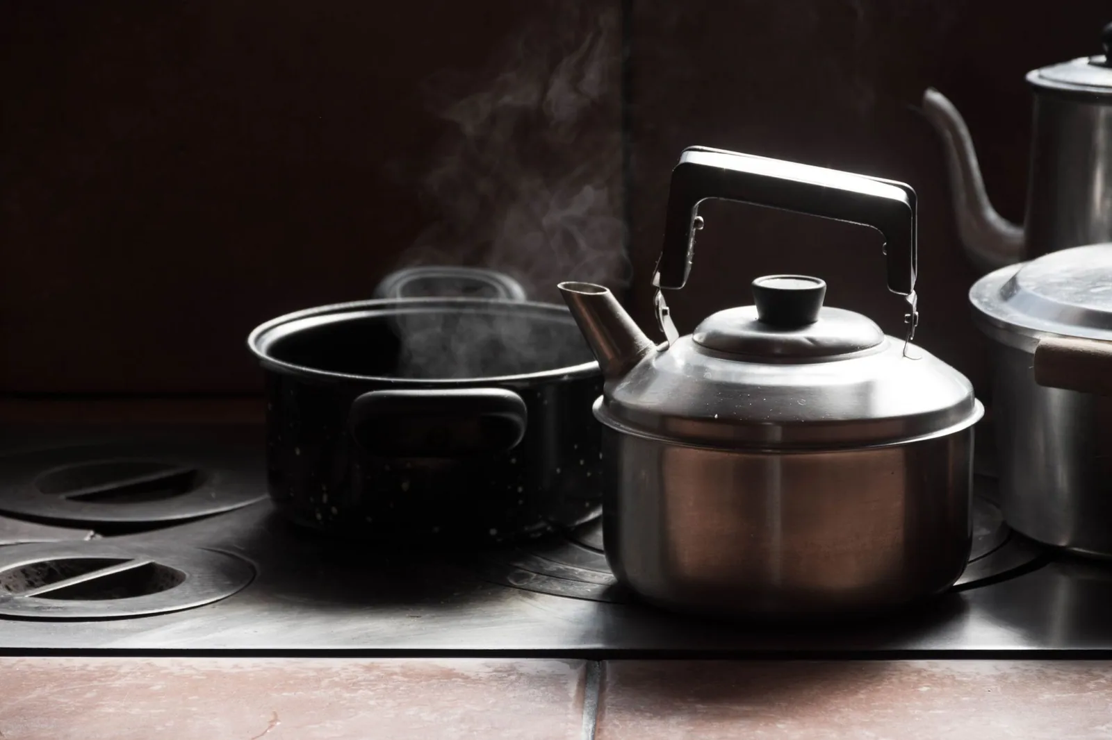 A metal kettle steaming on a kitchen stove beside a covered pot