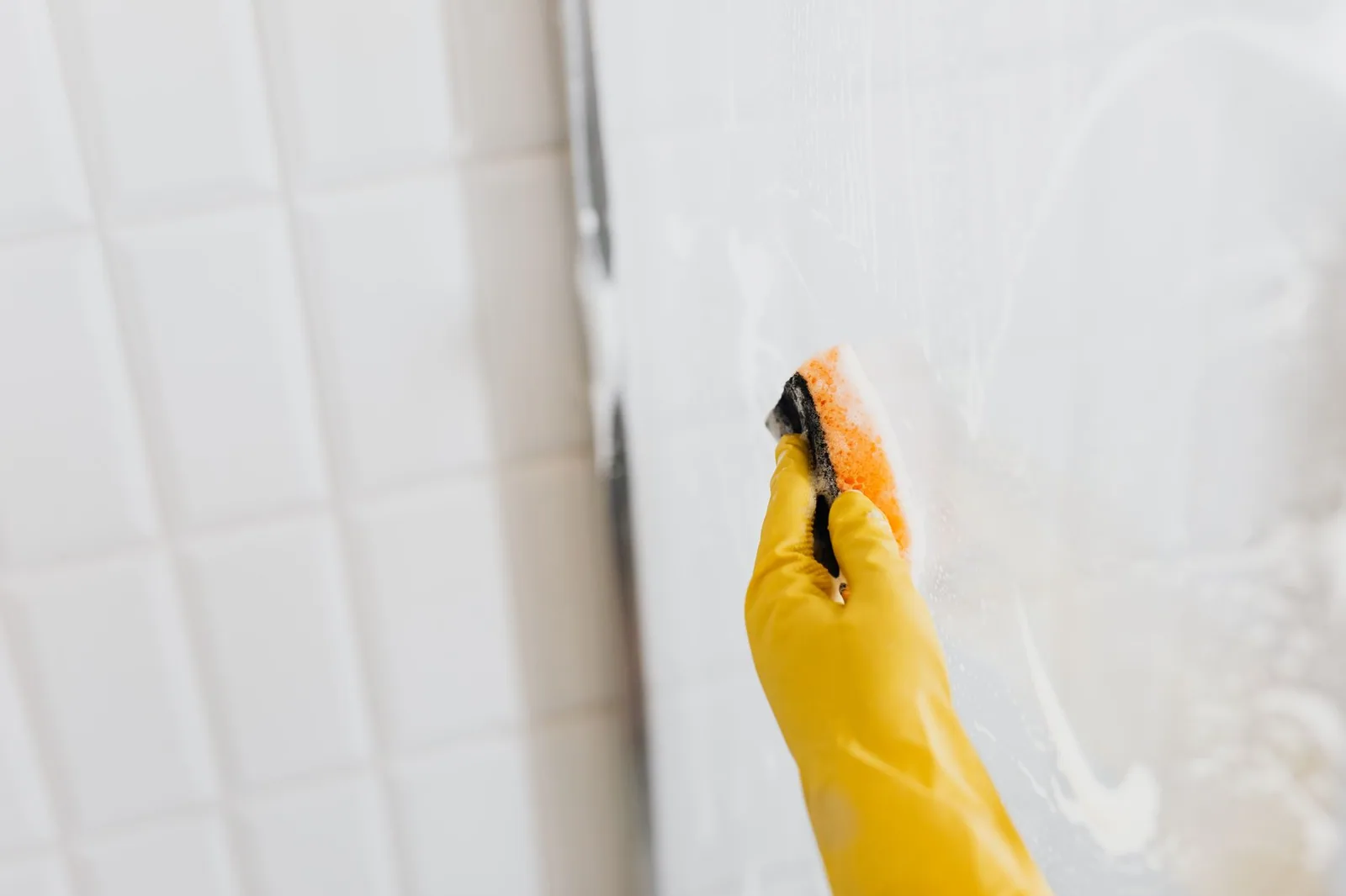 A gloved hand scrubbing a shower glass panel with a soapy sponge