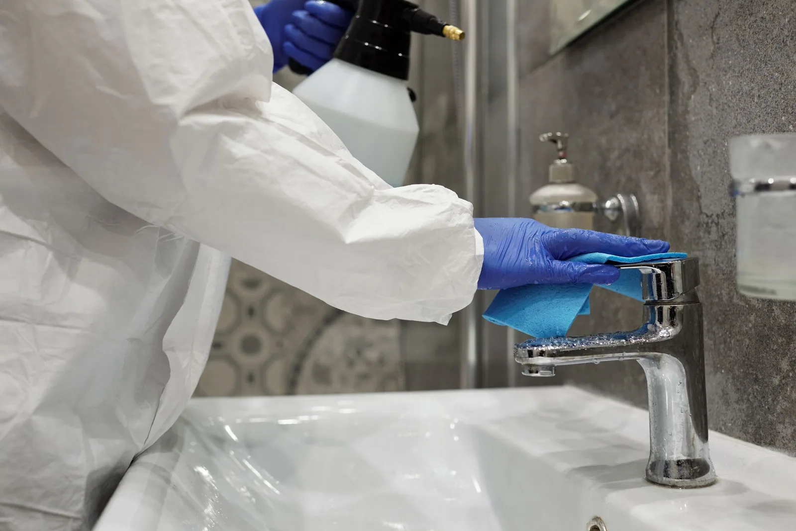 A gloved hand wiping a chrome bathroom faucet with a cloth and a spray bottle