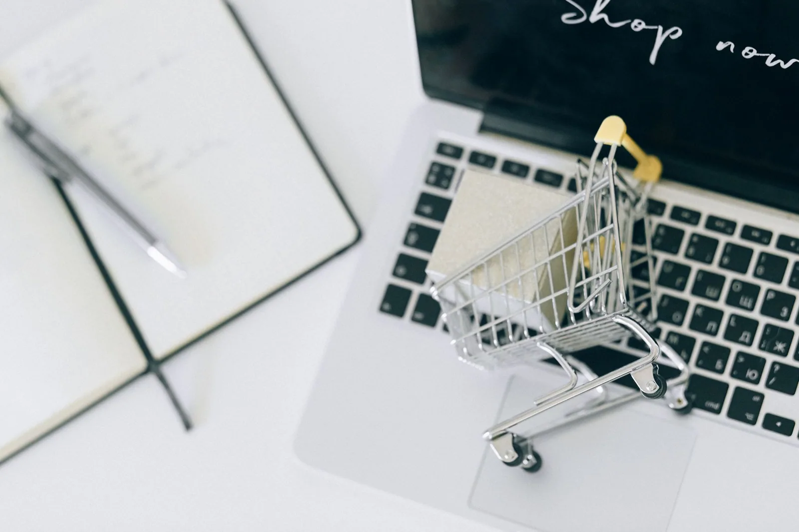 A miniature shopping cart on a laptop keyboard beside a notebook and pen