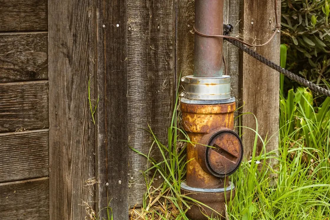 A rust-covered steel pipe fitting at the foot of a weathered wooden wall, surrounded by long grass