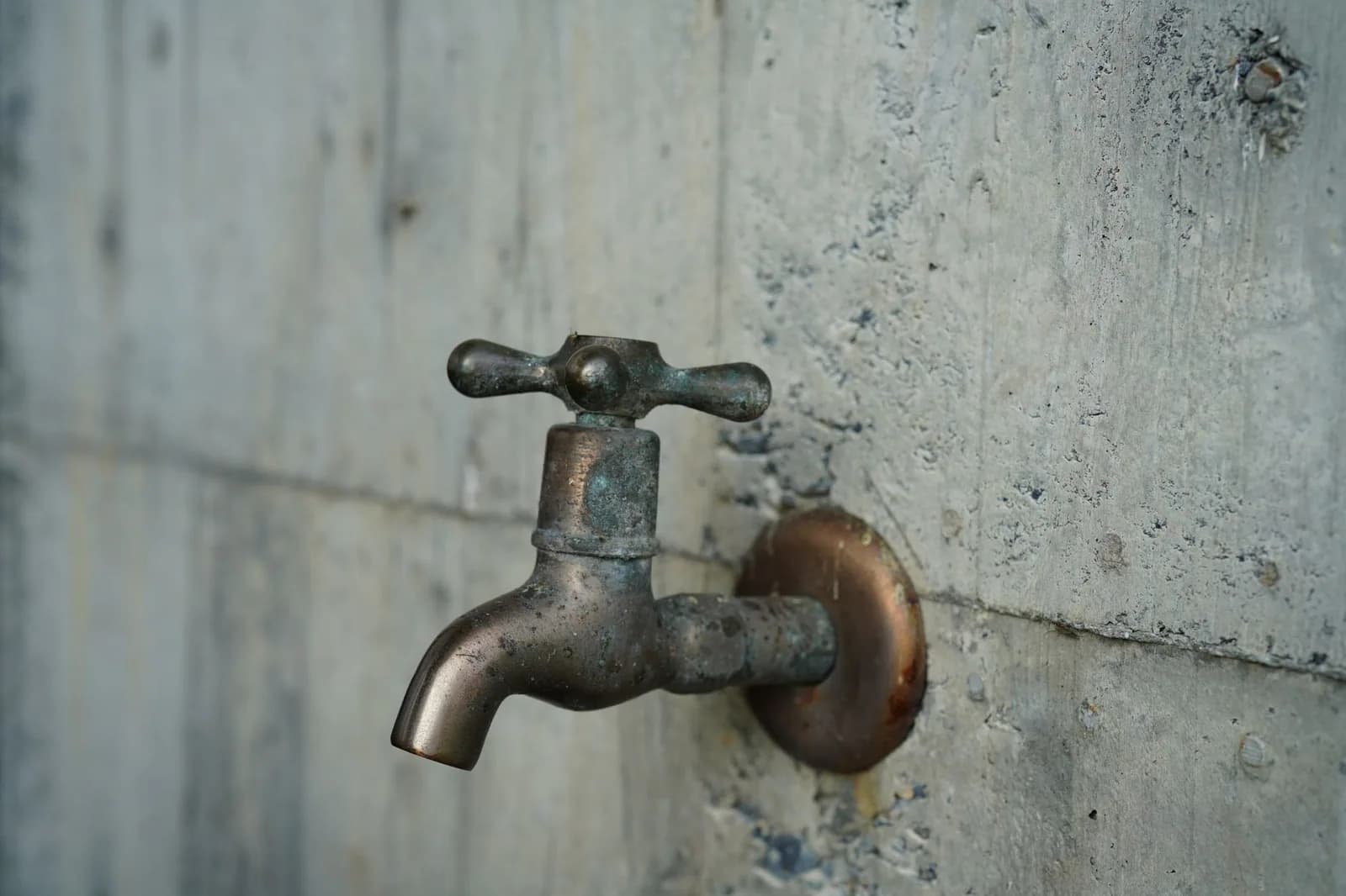 An old metal faucet on a concrete wall, its round wall plate streaked with rust
