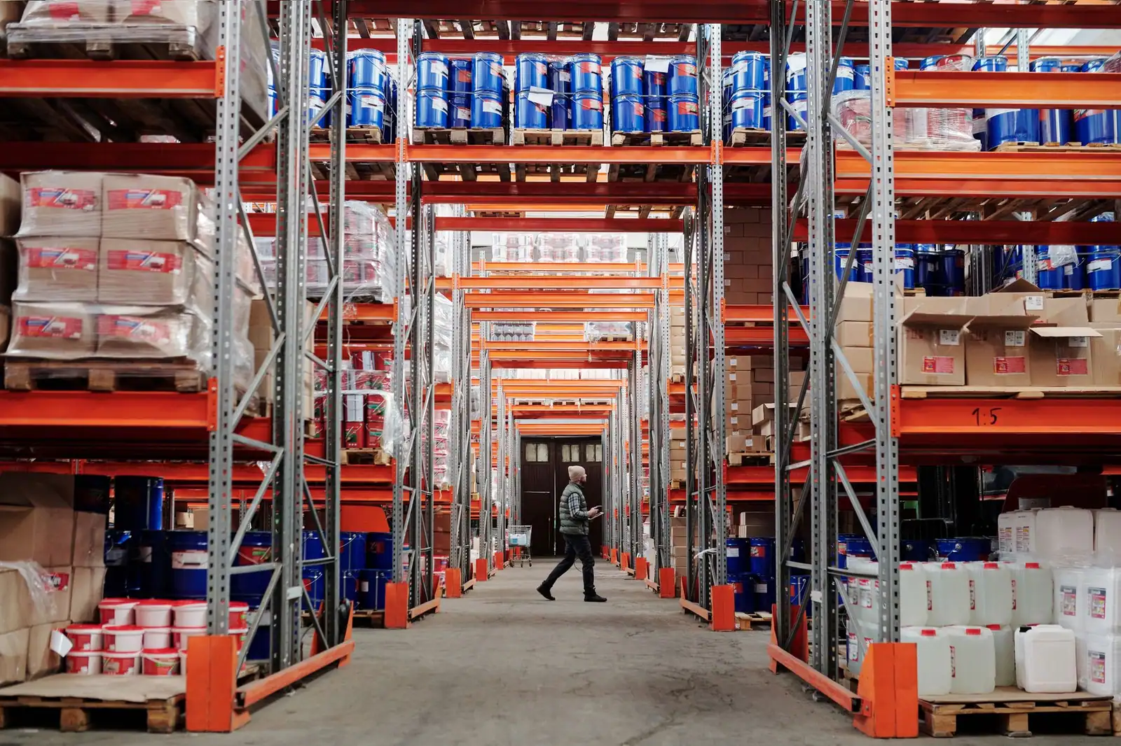 A worker walking a warehouse aisle stacked with boxed goods on pallet racking