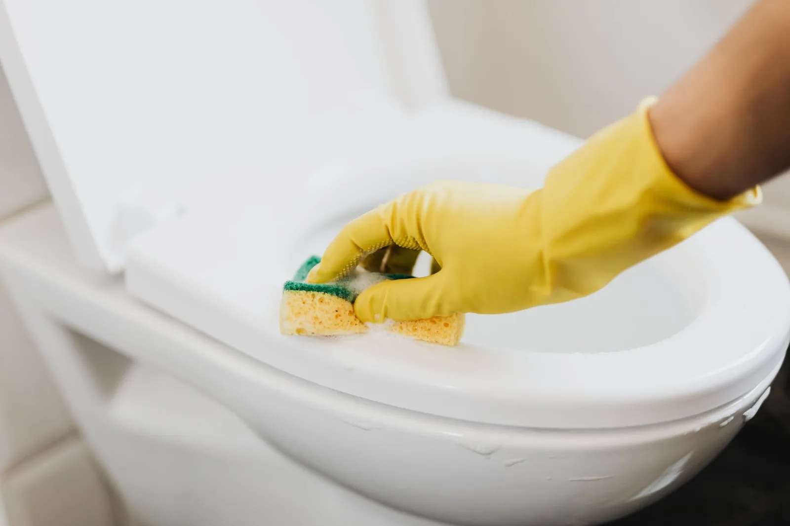 A gloved hand scrubbing a bathroom fixture with a soapy sponge